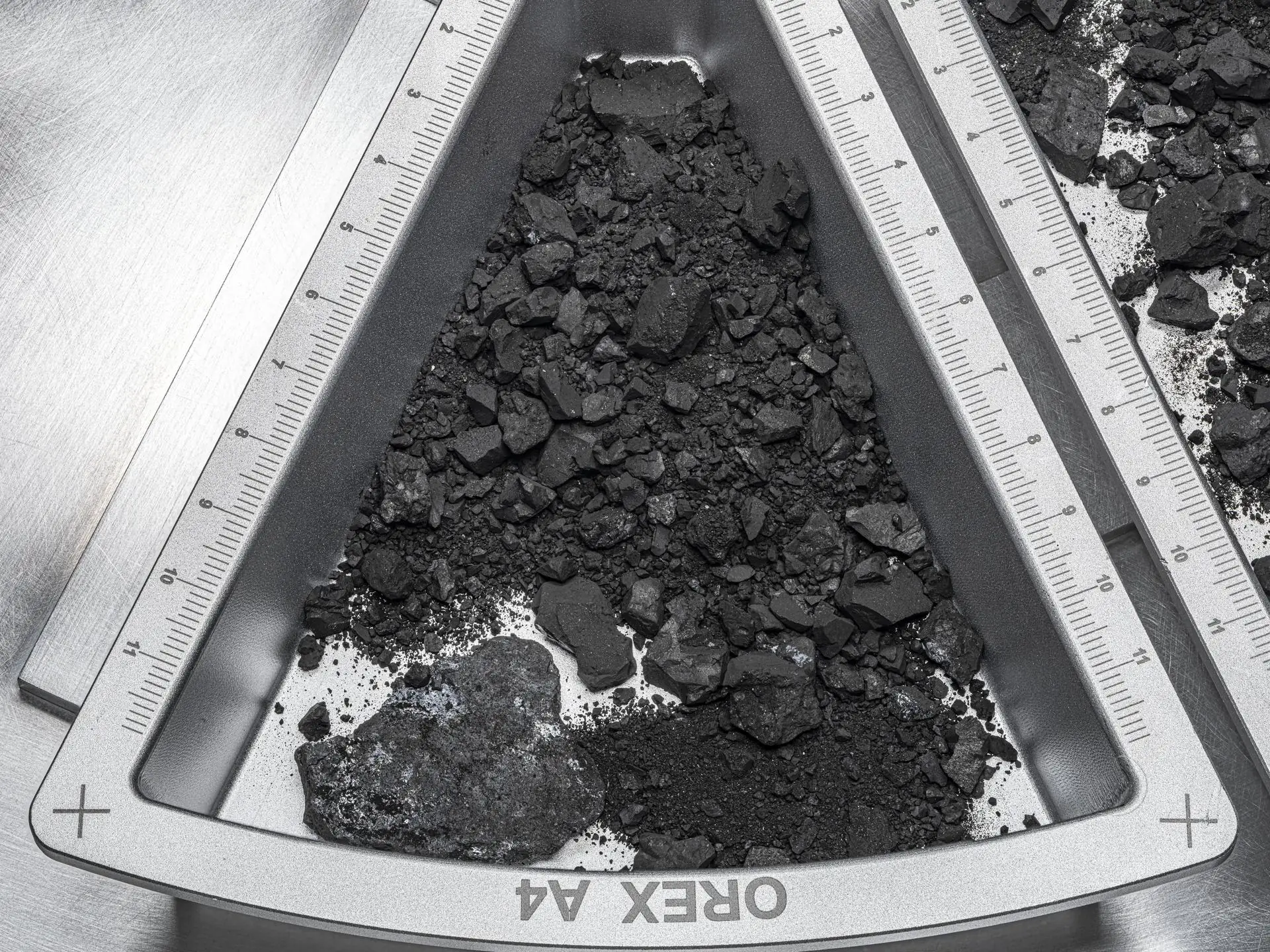 A triangular metal tray holding black rocks and dust from asteroid Bennu, with centimetre rulers along its edges