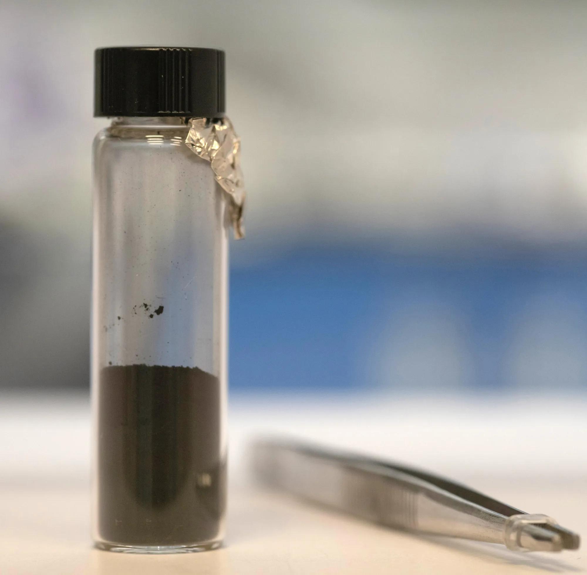 A glass vial with a black lid containing dark Bennu sample powder, with metal tweezers beside it