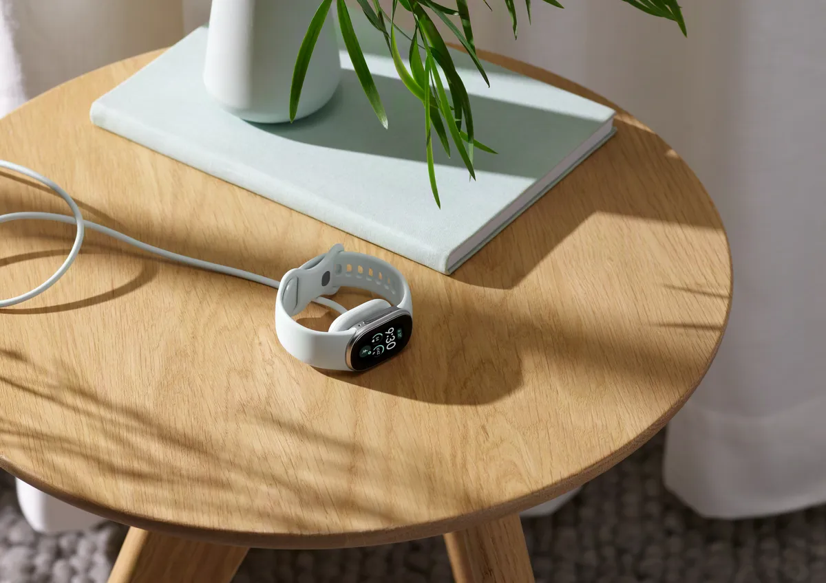 A Fitbit Edge with a white band charging on a wooden side table